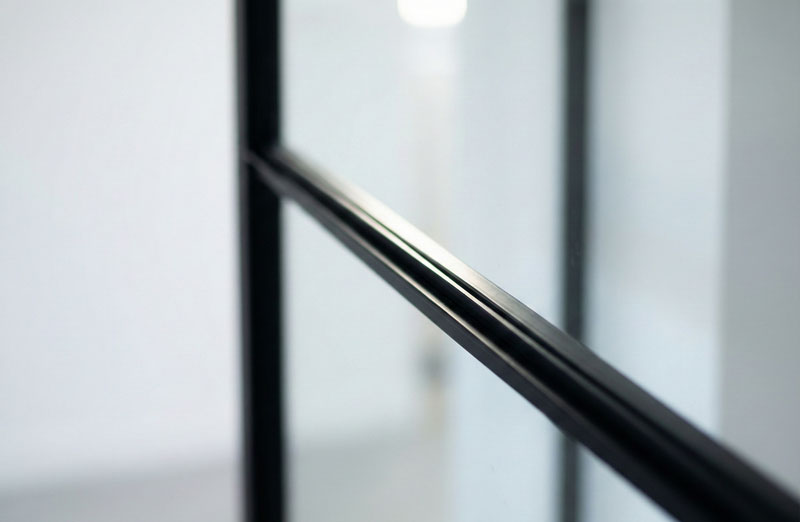 Un gros plan artistique en contre-plongée, centré sur le fin profilé en acier noir d'une cloison vitrée. La ligne métallique traverse l'image en diagonale, contrastant nettement avec le fond blanc et lumineux du mur et du plafond, qui sont légèrement floutés.